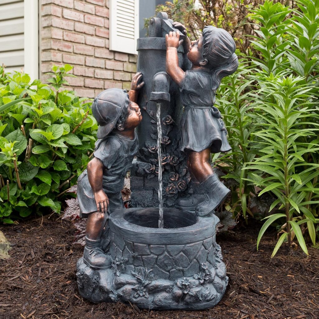 Whimsical small garden fountain statue of a boy and girl playing at a water pump surrounded by lush green plants and mulch.