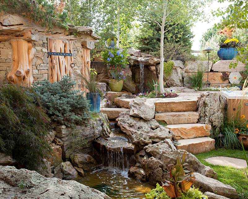 Rustic backyard garden with natural stone waterfall, rock steps, and lush landscaping plants.